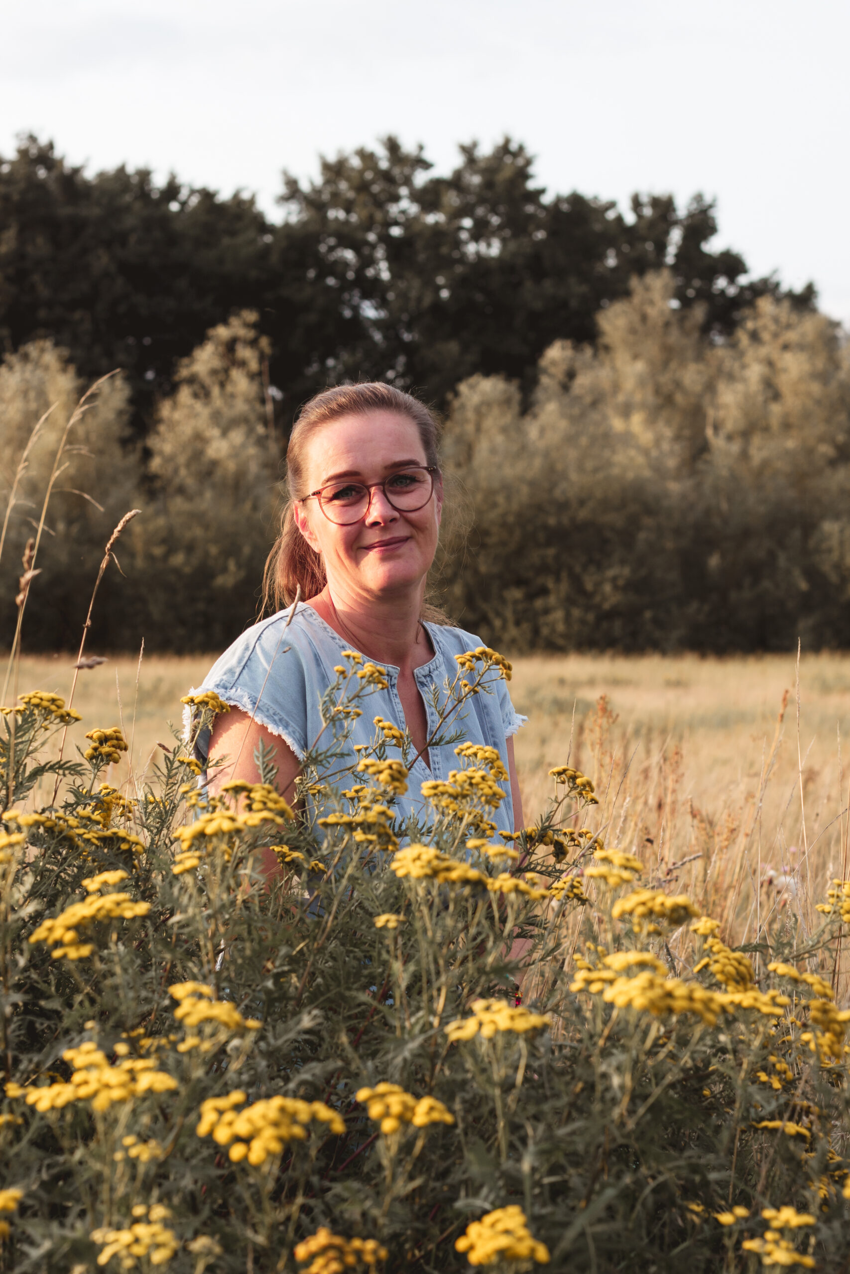 Auf dem Bild sieht man Jeannette Klemm. Sie sitzt hinter einem Blumenstrauch mit gelben Blüten. Im Hintergrund sieht man ein Getreidefeld was, an einem Wald grenzt. Man sieht Jeannette ab Brusthöhe. Sie trägt ein hellblaues Kleid was die Schultern bedeckt und die Arme frei lässt. Sie hat ihre Haare zu einem Pferdeschwanz gebunden und trägt eine Brille. Sie hat ihren Kopf leicht nach rechts geneigt und lächelt in die Kamera.
