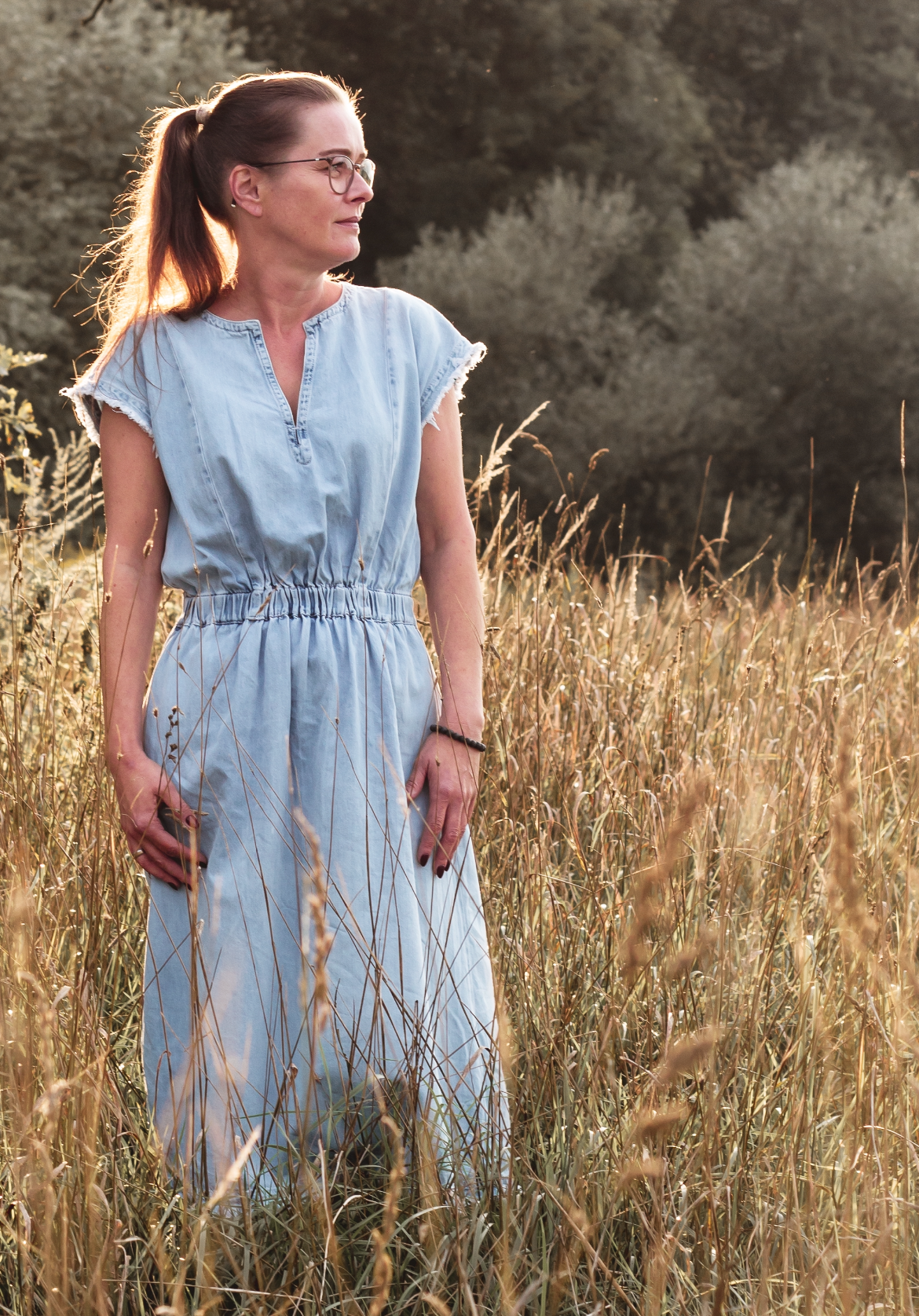Auf dem Bild sieht man Jeannette Klemm, wie sie auf einer Getreidewiese steht. Das Getreide hat die typische, sandige Farbe. Das Getreide geht ihr bis zur Hüfte. Sie hat ein blaues Kleid an, welches ihr bis zu den Knien reicht und nur die Schultern bedeckt. Ihre Arme hängen locker an ihrer Seite. Ihre Haare trägt sie zu einem Pferdeschwanz. Sie schaut verträumt nach links und trägt eine Brille.